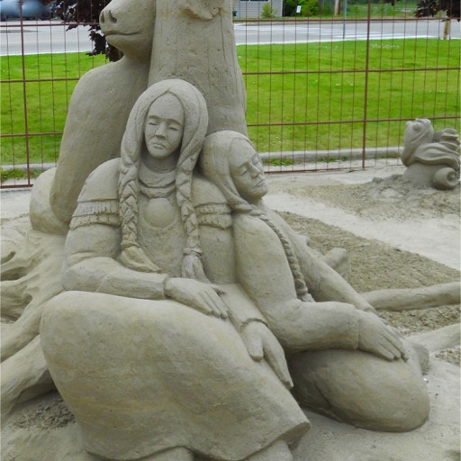 Sand sculpture of two princesses, seated and leaning against each other and a tree, sleeping soundly. The bear on the other side of the tree is watching over them. 