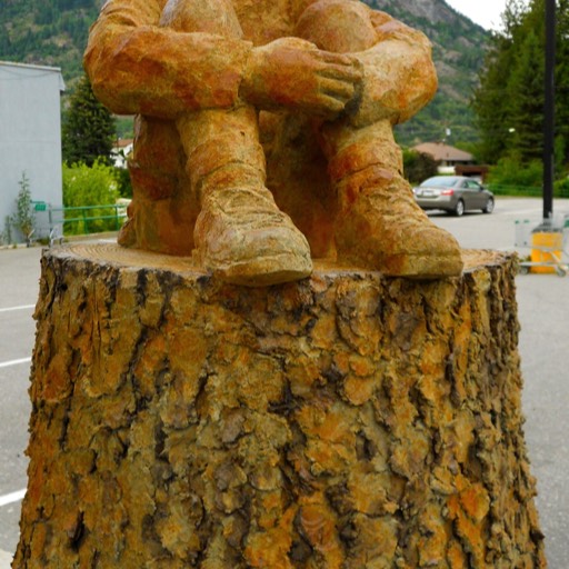 Wood sculpture of a boy sitting on a tree stump with his arms clasped around his shins. He is looking up and dreaming. 