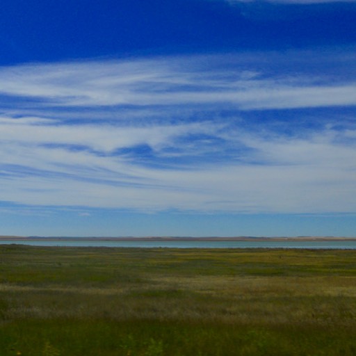 The sky is blue with a few thin swirling clouds. The prairie stretches, and a body of water can be seen in the distance. 