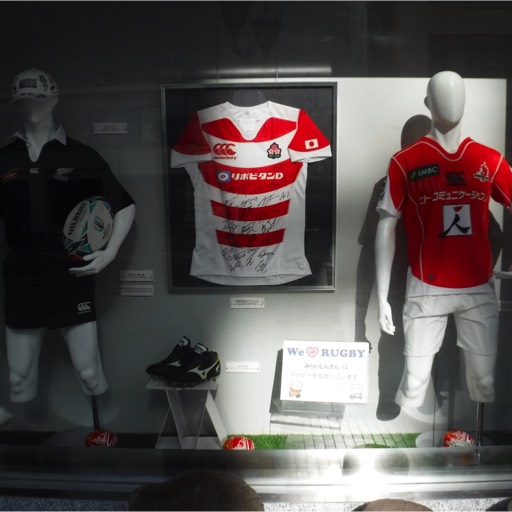 A shop window display with an autographed Japan rugby jersey in the centre. On each side is a mannequin. The mannequin on the left is cradling a rugby ball in its left arm and is dressed as a New Zealand All Black. The mannequin on the right is dressed in the national rugby uniform of Japan. 