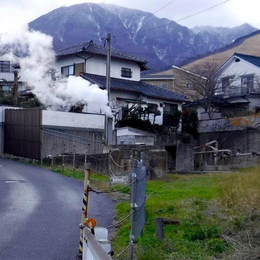 Houses in the foreground and a mountain in the background. In the middle of the frame is a pipe, embedded in the ground, from which a significant amount of steam is rising. 