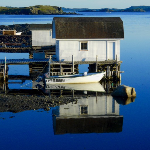 Photo of a boathouse with a small boat in front. The boat and house are in a bay, with ocean and land in the background. The boat house and boat are reflected on the still water.